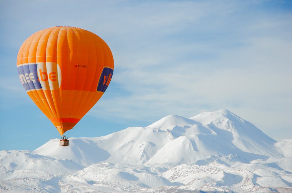 Orange hot air balloon flying over snow-covered mountains under a clear blue sky.